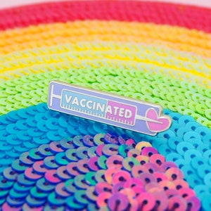 Puede incluir: Un pin de esmalte plateado y azul con forma de jeringa con la palabra "VACCINATED" en él. El pin está sobre un fondo de lentejuelas arcoíris.