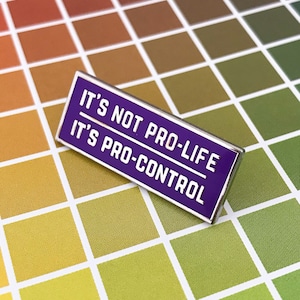 Puede incluir: Pin de esmalte morado con el texto "It's not pro-life, it's pro-control" en letras blancas. El pin está sobre un fondo blanco con una cuadrícula de cuadrados de colores.