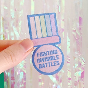Puede incluir: Un parche bordado azul y rosa con un sombrero a rayas rosa y blanco y el texto "Fighting Invisible Battles".
