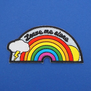 Puede incluir: Un parche bordado con forma de arcoíris con una nube blanca y un rayo en el lado izquierdo y un sol amarillo en el lado derecho. El texto "Leave me alone" está bordado en negro en la parte superior del arcoíris.