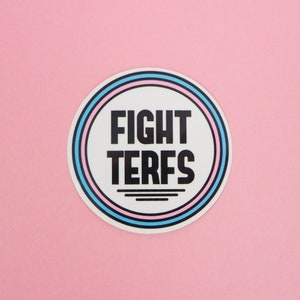 May include: A white circle sticker with a blue and pink ring around it. The text "FIGHT TERFS" is printed in black letters inside the circle.