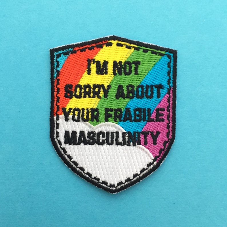 以下が含まれることがあります： 虹色のデザインと「I'M NOT SORRY ABOUT YOUR FRAGILE MASCULINITY」の文字が入った、シールド型の刺繍パッチ。 パッチは黒い縁取りがあり、水色の背景に配置されています。