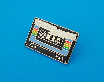 May include: A black and blue cassette tape pin with gold trim. The tape has a rainbow stripe and the text "Beethoven Best of Queen".