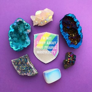 Shield-shaped embroidered patch that reads I'm Not Sorry About Your Fragile Masculinity in white capitals. Behind the words is a pastel rainbow and cloud. The patch has a white merrowed edge. The patch is surrounded by colourful crystals.