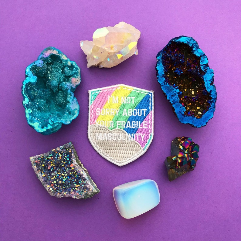 Shield-shaped embroidered patch that reads I'm Not Sorry About Your Fragile Masculinity in white capitals. Behind the words is a pastel rainbow and cloud. The patch has a white merrowed edge. The patch is surrounded by colourful crystals.