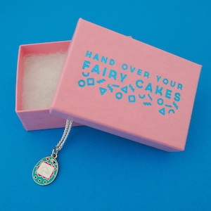 Silver-plated curb chain necklace with an enamel charm pendant. Charm is a teal, egg-shaped virtual pet toy. Necklace is shown emerging from a pink branded box.