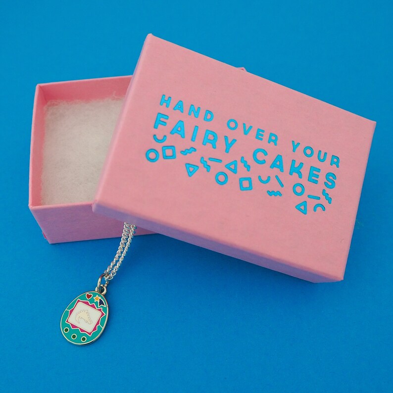 Silver-plated curb chain necklace with an enamel charm pendant. Charm is a teal, egg-shaped virtual pet toy. Necklace is shown emerging from a pink branded box.