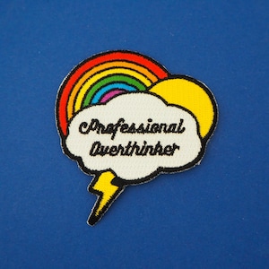 May include: A white embroidered patch with a rainbow, a yellow sun, and a speech bubble that says "Professional Overthinker".
