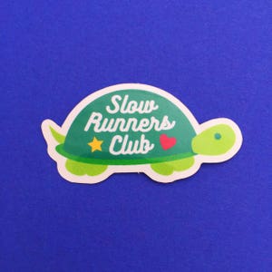 以下が含まれることがあります： 「Slow Runners Club」の白文字が入った、緑色のカメの形をしたステッカー。カメの甲羅は緑色で、黄色い星と赤いハートが付いています。背景は単色の青色です。
