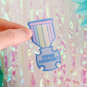 May include: A blue and pink felt patch in the shape of a medal with the text "Coping Admirably" on the front.