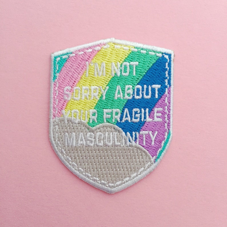Shield-shaped embroidered patch that reads I'm Not Sorry About Your Fragile Masculinity in white capitals. Behind the words is a pastel rainbow and cloud. The patch has a white merrowed edge.