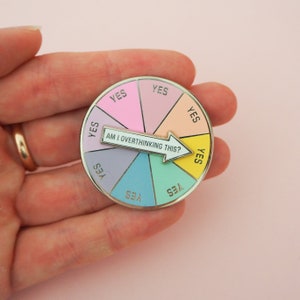 Circular pin divided into eight pastel coloured sections. Each section has the word Yes in capital letters. There is a white arrow that can spin to point to any section with the words Am I Overthinking This? The pin is shown on a hand for scale.