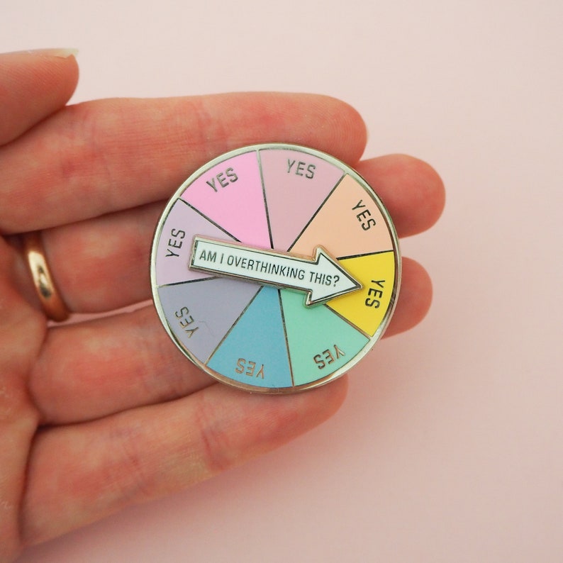 Circular pin divided into eight pastel coloured sections. Each section has the word Yes in capital letters. There is a white arrow that can spin to point to any section with the words Am I Overthinking This? The pin is shown on a hand for scale.