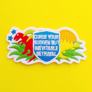 May include: White embroidered patch with a blue shield featuring the text "Curse your sudden but inevitable betrayal". The shield is surrounded by green grass, a red dinosaur, and a yellow dinosaur.