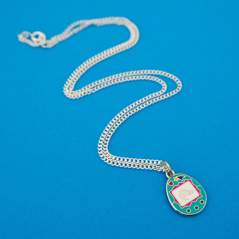 Silver-plated curb chain necklace with an enamel charm pendant. Charm is a teal, egg-shaped virtual pet toy. On the screen is a silver stegosaur & game icons. Beneath the screen are 3 buttons and above are a heart and shooting star in bright colours.