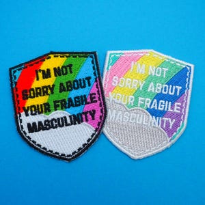 May include: Two embroidered patches in a shield shape. One patch has a black border and a rainbow design with the text "I'M NOT SORRY ABOUT YOUR FRAGILE MASCULINITY". The other patch has a white border and a similar design.