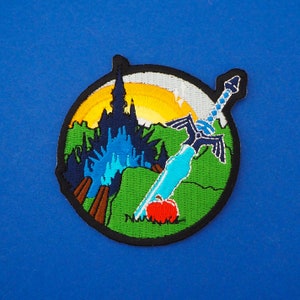 May include: A colorful embroidered patch featuring a blue and silver sword, a castle, and an apple. The patch is set against a green background with a sunset in the background.
