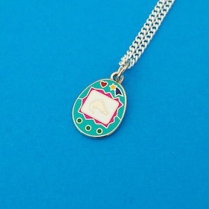 Silver-plated curb chain necklace with an enamel charm pendant. Charm is a teal, egg-shaped virtual pet toy. On the screen is a silver stegosaur & game icons. Beneath the screen are 3 buttons and above are a heart and shooting star in bright colours.