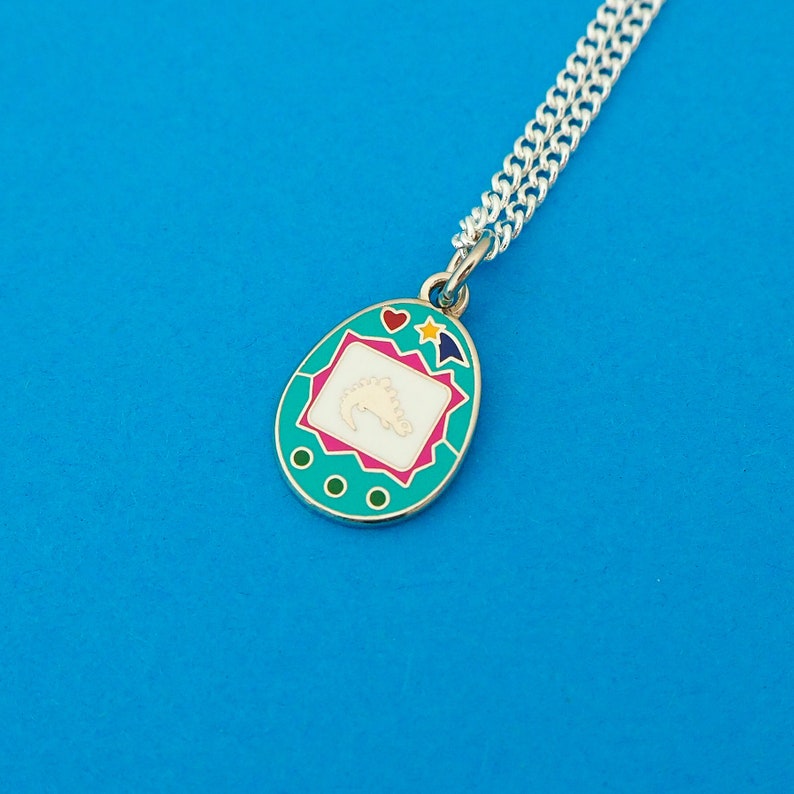Silver-plated curb chain necklace with an enamel charm pendant. Charm is a teal, egg-shaped virtual pet toy. On the screen is a silver stegosaur & game icons. Beneath the screen are 3 buttons and above are a heart and shooting star in bright colours.
