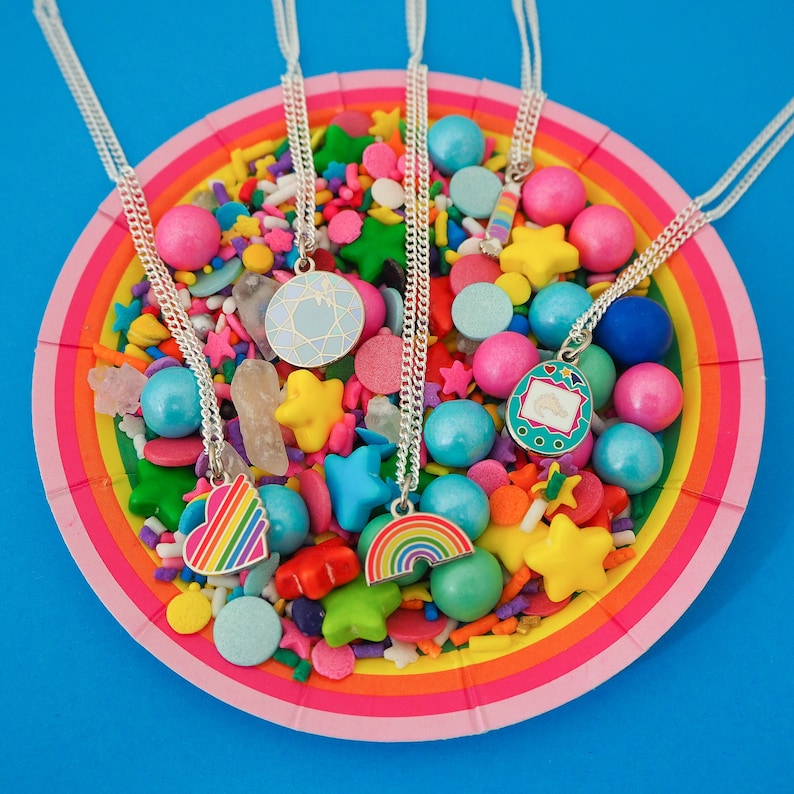 Five silver curb-chain necklaces, each with an enamel charm pendant. The pendants are of different colourful designs. The necklaces are draped over a plate full of colourful candy.