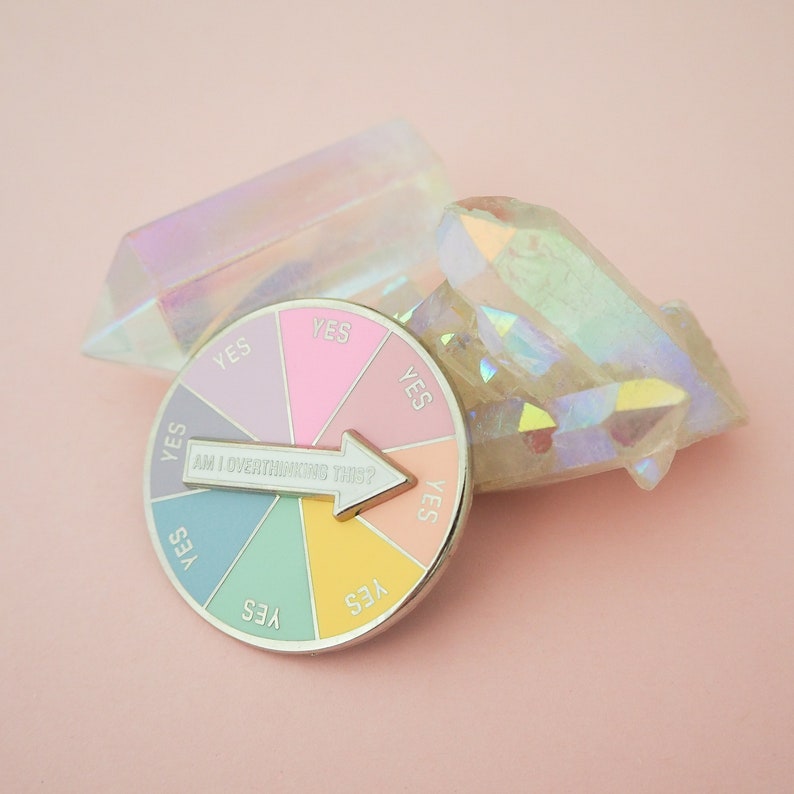 Circular pin divided into eight pastel coloured sections. Each section has the word Yes in capital letters. There is a white arrow that can spin to point to any section with the words Am I Overthinking This? The pin is leaning on a quartz crystal.
