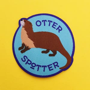 Peut inclure: Un écusson brodé bleu et violet avec une loutre brune sur un fond bleu clair. Le texte "Otter Spotter" est brodé en violet.