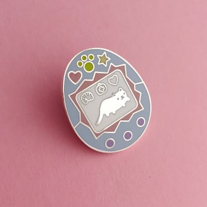 May include: Enamel pin in the shape of a retro handheld game, featuring a white cat illustration. The pin has a light blue border with pink and white accents, and includes paw prints, hearts, and a star design. The pin is set against a pink background.