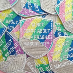 Scattered pile of shield-shaped embroidered patches that read I'm Not Sorry About Your Fragile Masculinity in white capitals. Behind the words is a pastel rainbow and cloud. The patch has a white merrowed edge.