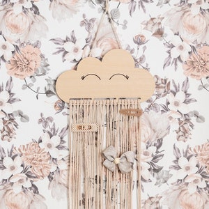 May include: A wooden cloud-shaped hair accessory holder with a smiling face. The holder has a long, beige macrame fringe hanging from the bottom. A bow and hair clips are displayed on the fringe.