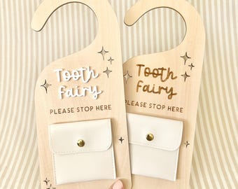 May include: Two wooden door hangers with a cutout design, each featuring the text "Tooth Fairy" and a small pocket. The text "PLEASE STOP HERE" is also included. The hangers are light-colored with a cream-colored pocket and gold button.