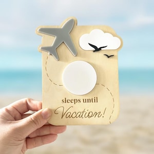 May include: A wooden vacation countdown sign with a gray airplane, white cloud, and black birds. The sign has a white circle and the text "sleeps until Vacation!" in a script font. The background is a beach scene.