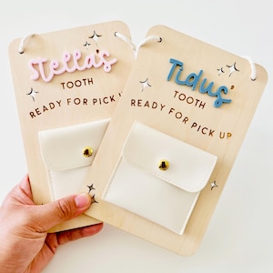 May include: Two wooden tooth fairy boards with the names "Stella's" and "Tidus" in pink and blue, respectively. Each board has a small white pouch with a gold button closure. The text "TOOTH READY FOR PICK UP" is printed on each board.