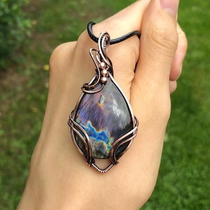May include: A teardrop-shaped labradorite pendant featuring iridescent blue and purple flashes, encased in a decorative copper wire-wrapped setting with intricate swirls and small metal beads, suspended from a black cord necklace.