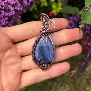 May include: A teardrop-shaped labradorite pendant featuring a polished blue stone encased in intricate copper wire wrapping. The metalwork includes coiled loops and decorative swirls that secure the gemstone within a structured frame.