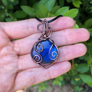 May include: A pendant featuring a polished, iridescent blue labradorite gemstone encased in intricate, hand-wrapped copper wire. The wirework forms decorative swirls and loops around the stone, suspended from a simple black cord necklace.