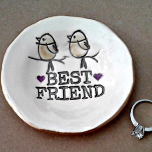 May include: White ceramic ring dish with a gold rim. Two birds are perched on a branch with the words "Best Friend" and two purple hearts.