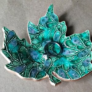 May include: A ceramic leaf-shaped dish with a green and blue swirling pattern. The dish has a gold rim and a ring is resting in the center.