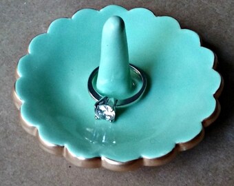 May include: A mint green ceramic ring dish with a gold rim and a small white ceramic ring holder in the center. A silver ring with a clear stone is on the holder.