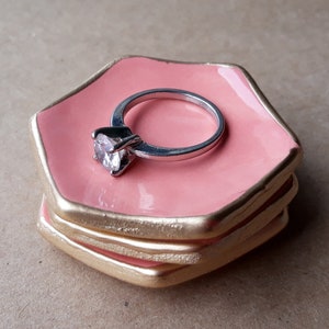 May include: A silver ring with a large, clear gemstone rests on a stack of three pink ceramic ring dishes. The dishes have gold-colored edges.
