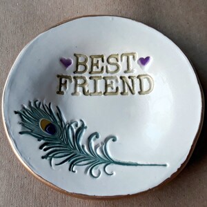 May include: White ceramic trinket dish with a gold rim. The dish has a peacock feather design and the words "BEST FRIEND" with two purple hearts.