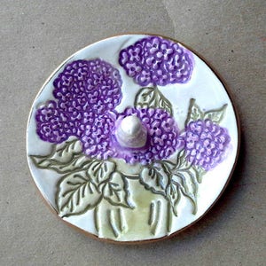 May include: A white ceramic ring dish with a floral design. The dish features three purple flowers with green leaves. The flowers are stylized and have a textured surface.