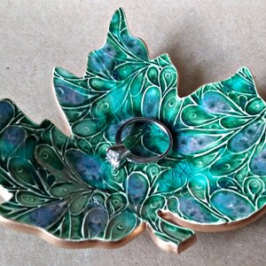 May include: A green ceramic ring dish shaped like a maple leaf with a swirling pattern. A silver ring with a clear stone is resting on the dish.
