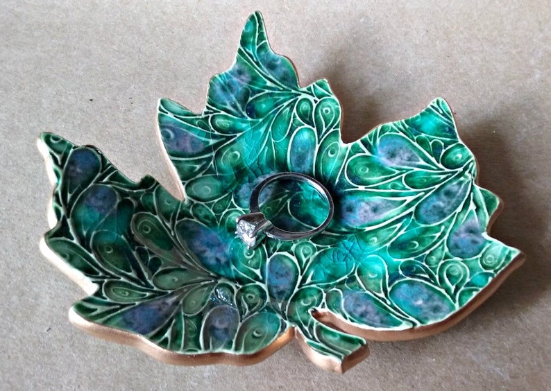 May include: A green ceramic ring dish shaped like a maple leaf with a swirling pattern. A silver ring with a clear stone is resting on the dish.