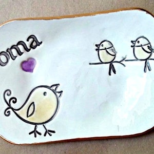 May include: White ceramic dish with a brown rim. The dish has a purple heart and the word "oma" written on it. There are three hand-drawn birds on the dish.