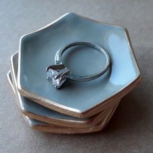 May include: A set of three gray ceramic ring dishes with gold trim. The top dish has a silver ring with a clear stone on it.