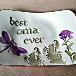 May include: White ceramic dish with the text "best oma ever" in green lettering. The dish is decorated with a purple dragonfly, a purple flower, and green leaves.