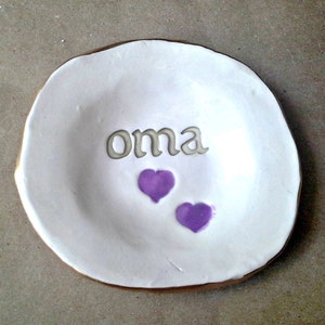 May include: White ceramic ring dish with a gold rim. The dish has the word "oma" written on it in a cursive font and two purple hearts.