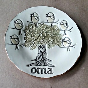May include: A white ceramic plate with a tree and birds painted on it. The tree has a heart in the trunk and the word "oma" is written below the tree.