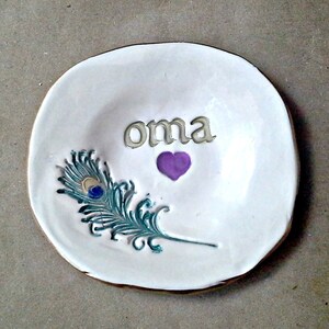 May include: White ceramic trinket dish with a gold rim. The dish has a peacock feather design and the word "oma" written in gold lettering. A purple heart is above the word.