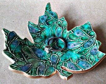 May include: A ceramic ring dish shaped like a green and blue maple leaf with intricate swirling patterns. A silver ring is resting in the center of the dish.